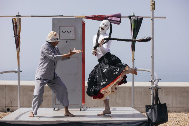 An image of a male and female performer stood on a make shift platform with a sunlit sky background.