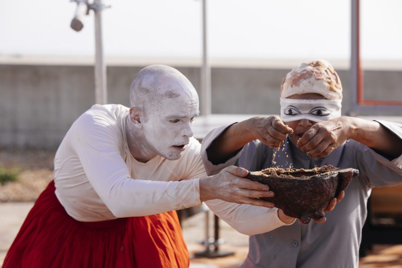 An image of a male and female performers. The male is showing her a pot of water from which she is washing her hands.