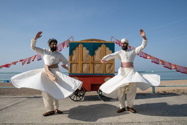 An image of two male Indian dancers stood in front of a wooden cart with bunting and a blue sky background.