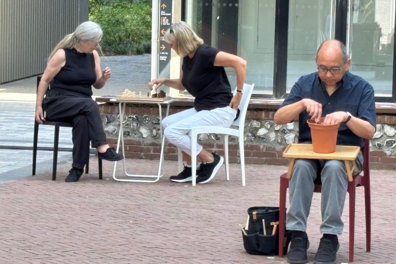 An image of two women and a man sat on some chairs around small tables. The two women are playing chess and the man is tending to a plant pot.
