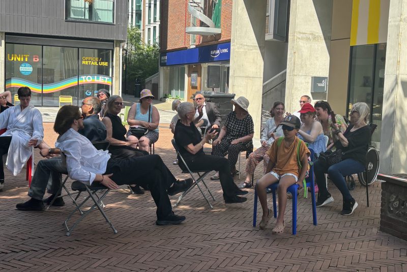 An image of a group of people sat on some chairs that are scattered around a courtyard area surrounded by buildings.