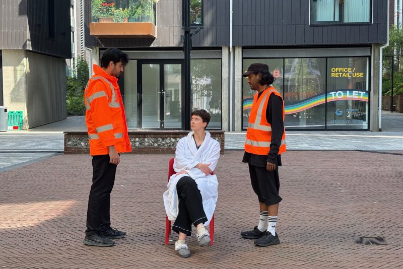 An image of two men stood facing a woman sat on a chair. Both men are wearing high vis orange jackets, and the woman is wearing a dressing gown and slippers.