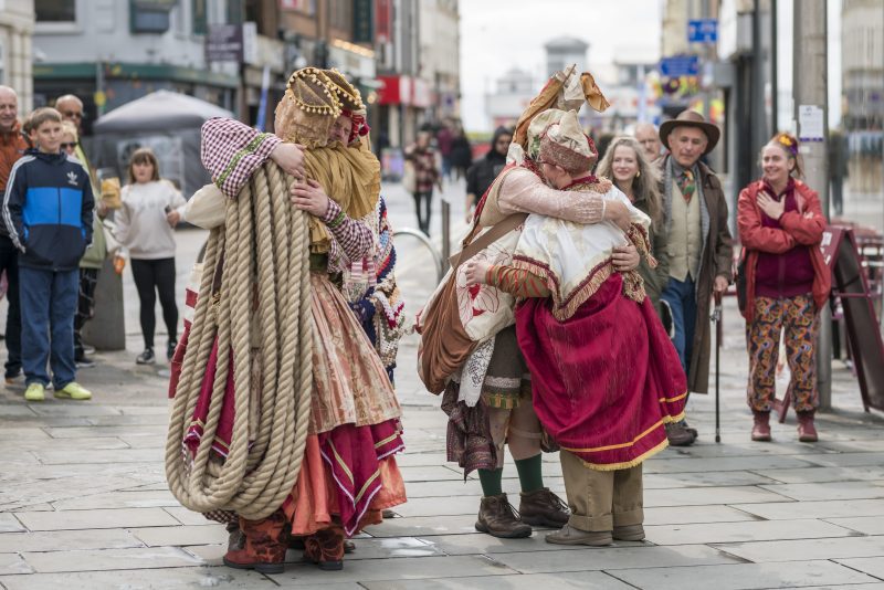 An image of a group of performers hugging in the street wearing regal clothing. One of them has a rope over their shoulder