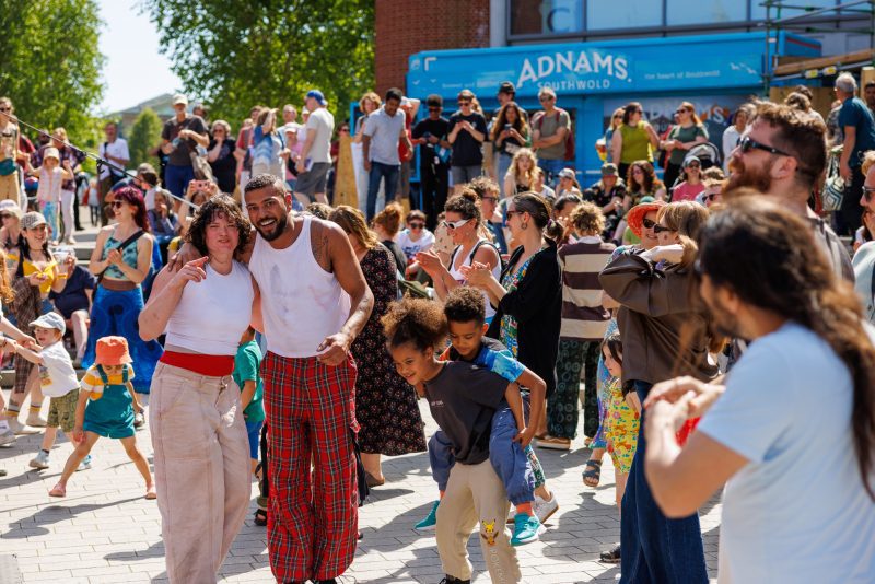 An image of a male and female performer with their arms around each other. There is a crowd of people stood behind them.