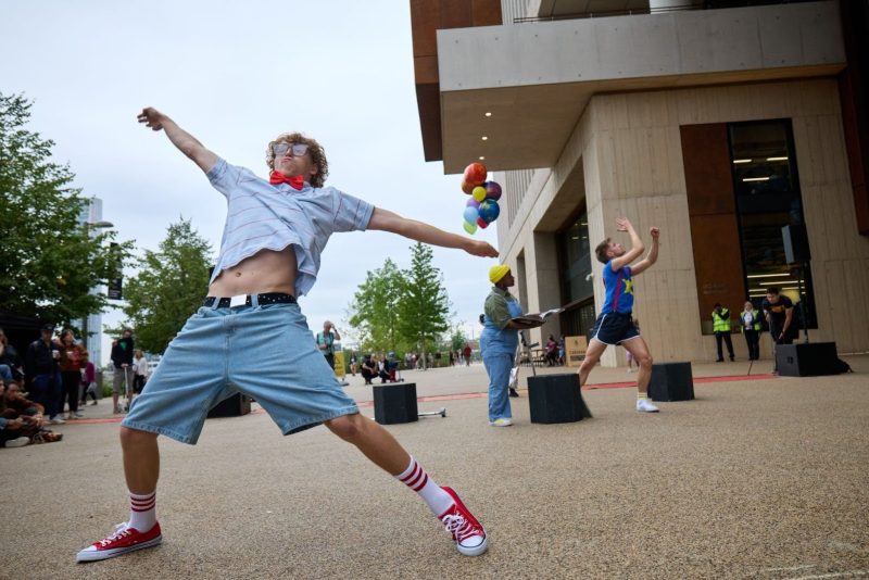 An image of three performers stood in front of a concrete building.