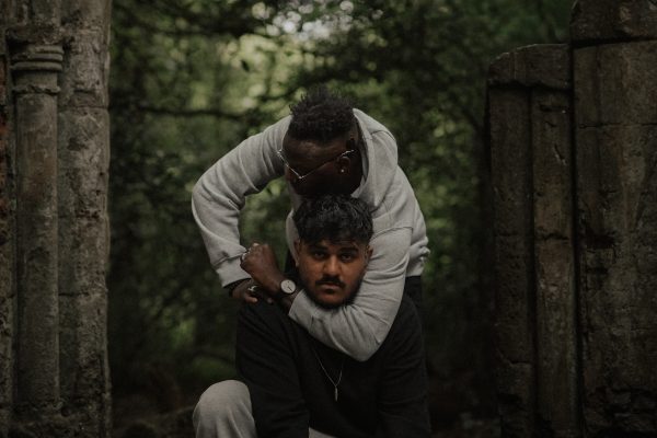 A portrait image of two men. One of them has the other's head in an arm lock, and there are trees in the background.