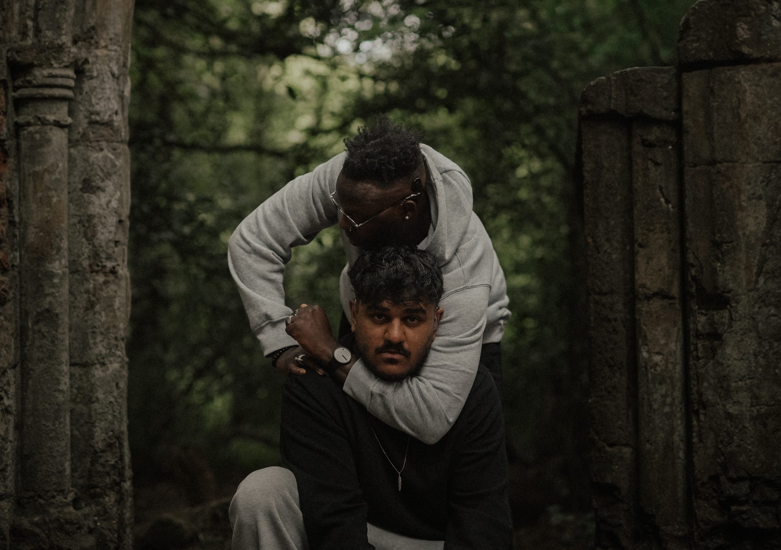 A portrait image of two men. One of them has the other's head in an arm lock, and there are trees in the background.