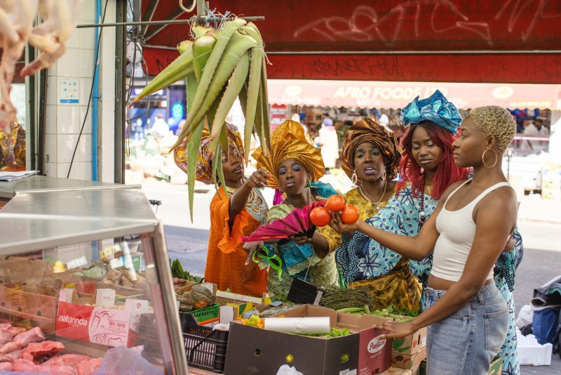 An image of five black women wearing traditional African dress and carrying oranges, stood in a fresh grocery store.