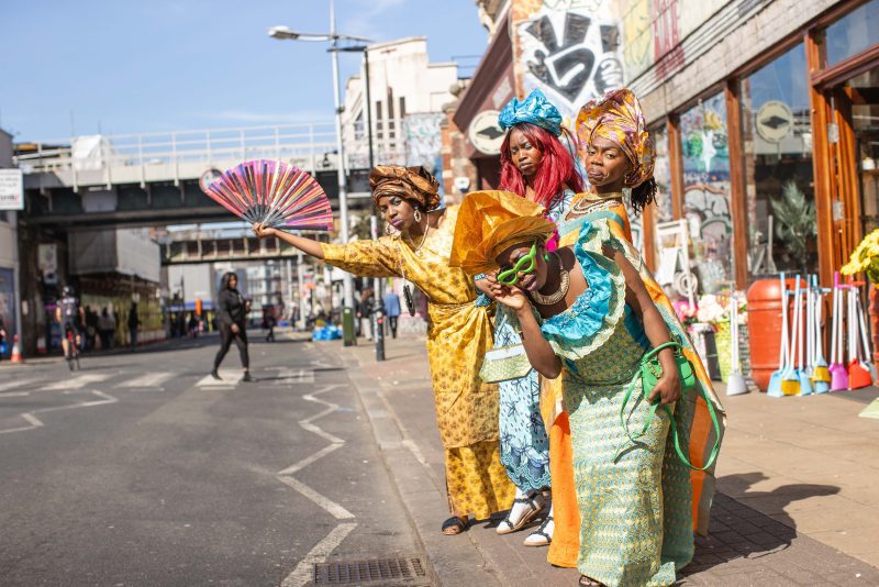 An image of five black women wearing traditional African dress stood at a London bus stop waiting for a bus.