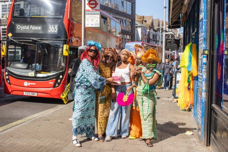 An image of five black women wearing traditional African dress stood at a bus stop in front of a London red bus