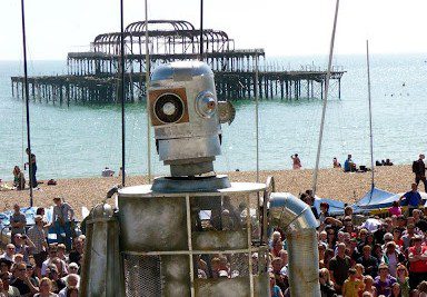 An image of a giant metallic figure with a small audience, pier, sea and beach in the background.