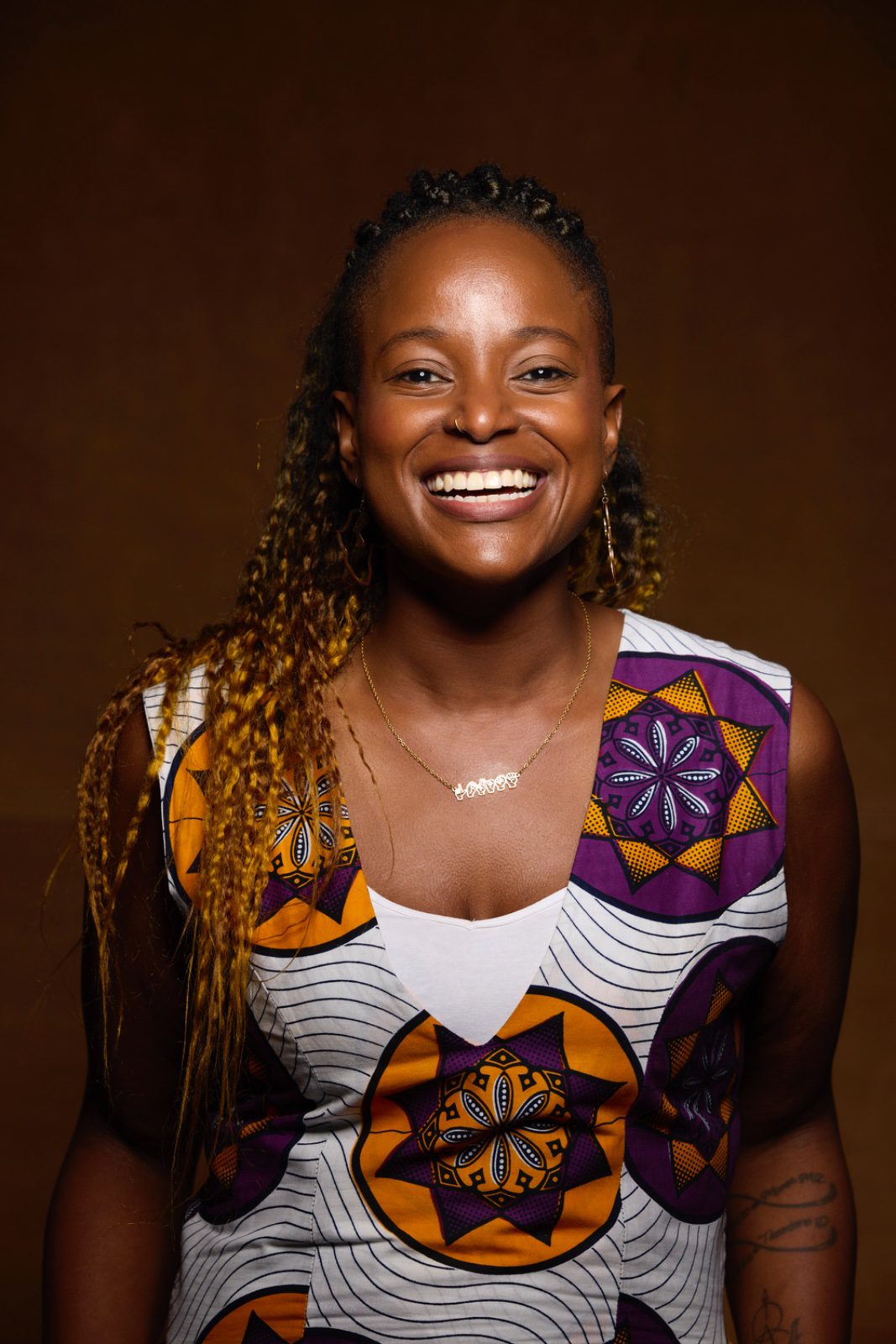 A portrait image of a black woman smiling at the camera wearing a colourful top with a plain brown background.