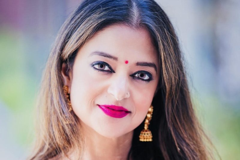 A portrait image of a woman with long dark hair, gold earrings and bright pink lipstick.