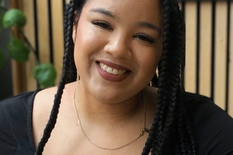 A portrait image of a mixed-race woman with long dark dreadlocks and wearing a black top.