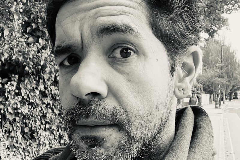 A black and white portrait image of a white man with short textured hair and a beard.
